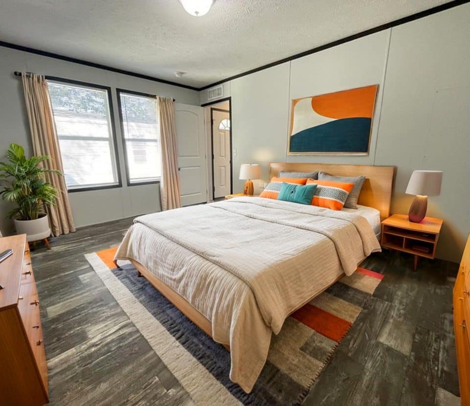 A modern bedroom with a large bed, neutral bedding, and colorful accent pillows. There are wooden nightstands, a dresser, a potted plant, large windows with curtains, and abstract wall art above the bed.