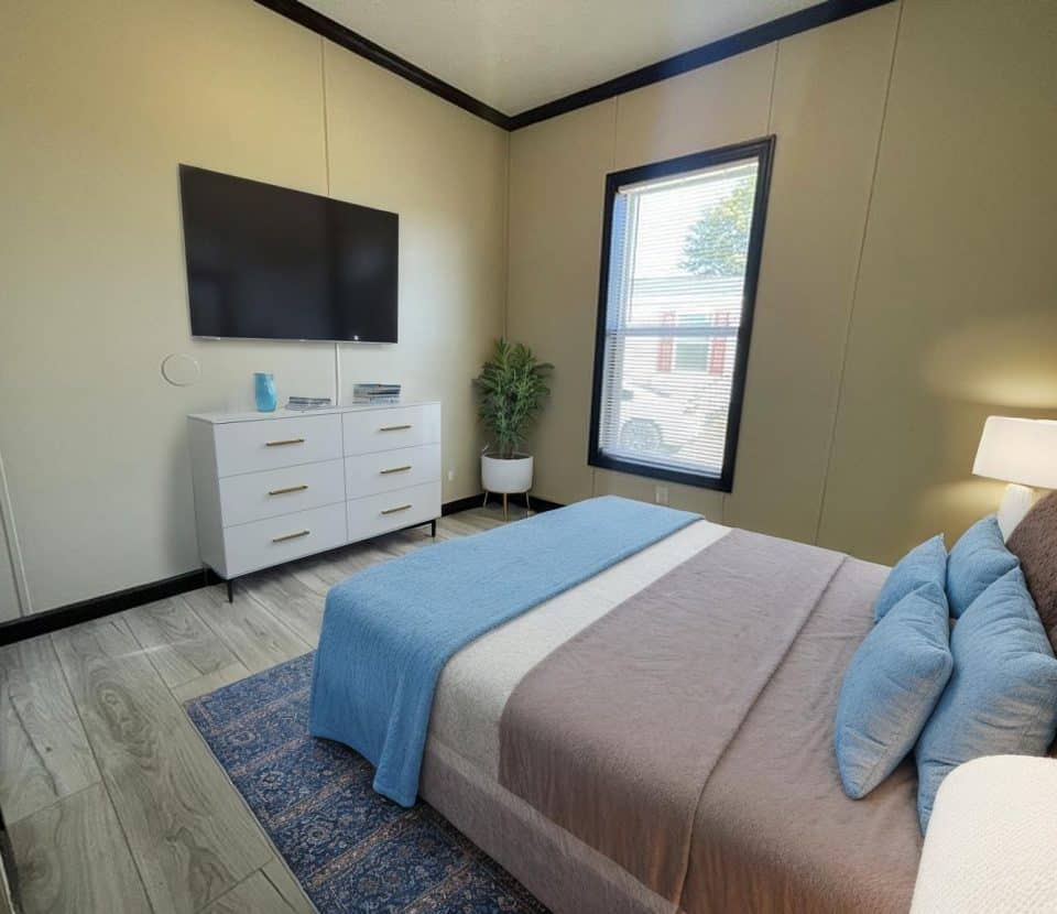 A cozy bedroom with a neatly made bed in blue and brown tones, a white dresser with decor, a large wall-mounted TV, a potted plant by the window, light wood flooring, and a rug. Natural light comes in through the window.
