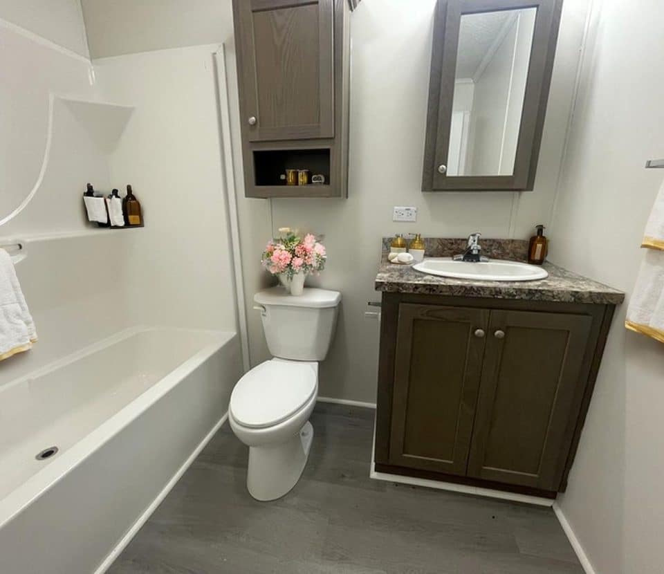 A clean, modern bathroom with a white bathtub, toilet, and sink. Brown cabinets and countertop, flowers on the toilet, towels, soap bottles, and a wall mirror create a tidy and inviting space.