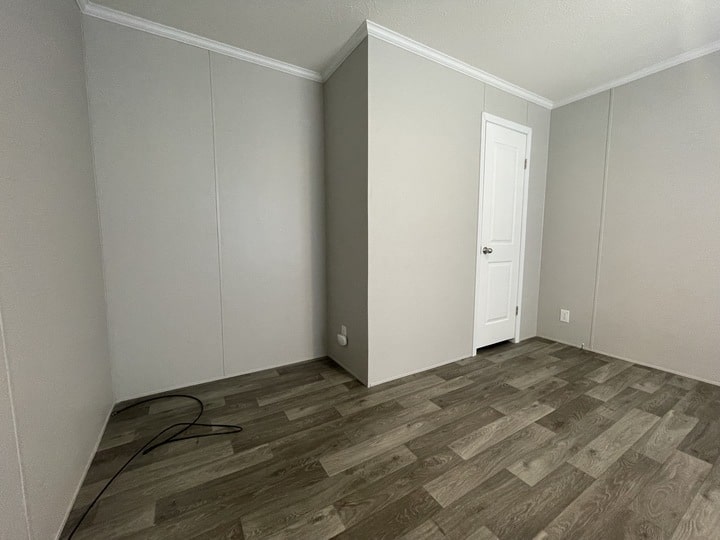 Empty room with light gray walls, wood-patterned vinyl flooring, a white door, and a few electrical outlets. A loose black cable is on the floor in the corner of the room.