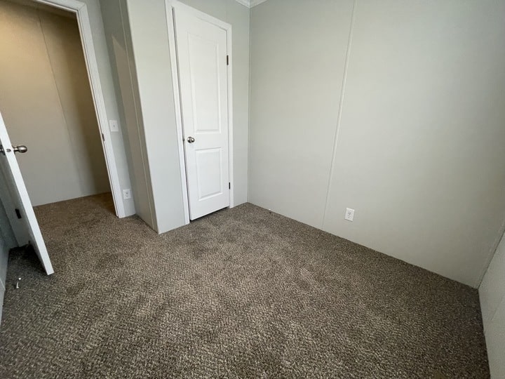 Small empty room with beige carpet, light gray walls, two white doors, and an electrical outlet on the right wall. One door is open, revealing a hallway with similar carpeting.