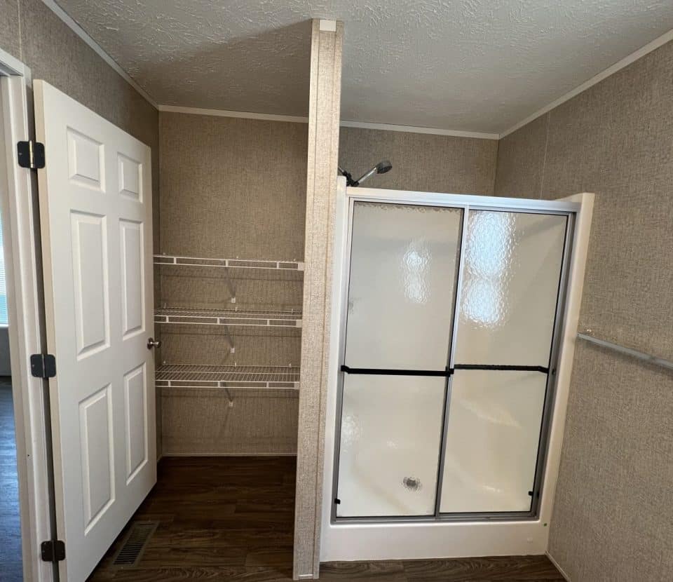 A bathroom with a white door, built-in wire shelves, a corner shower with glass sliding doors, beige textured walls, and dark wood flooring. Natural light enters from an adjacent room.
