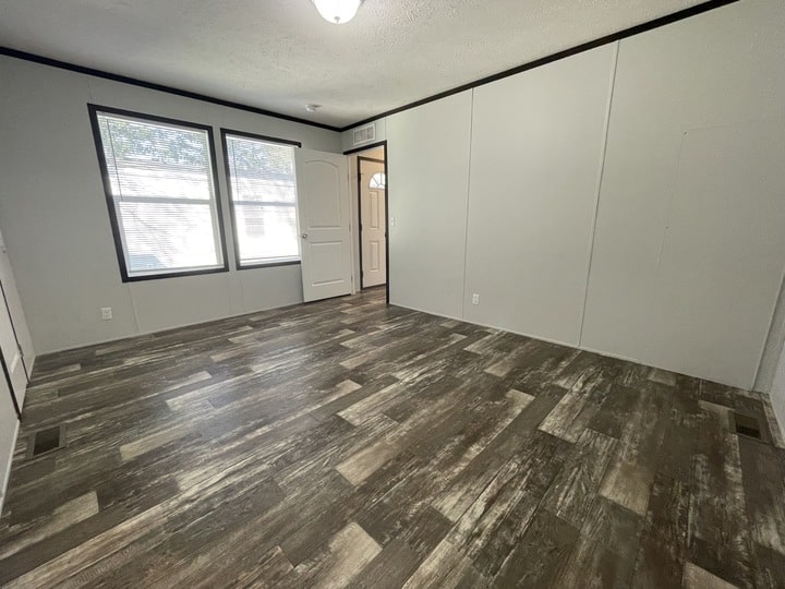 An empty room with wood-patterned vinyl flooring, white walls, two large windows with blinds, and a white door. Natural light streams in, giving the space a bright and open feel.