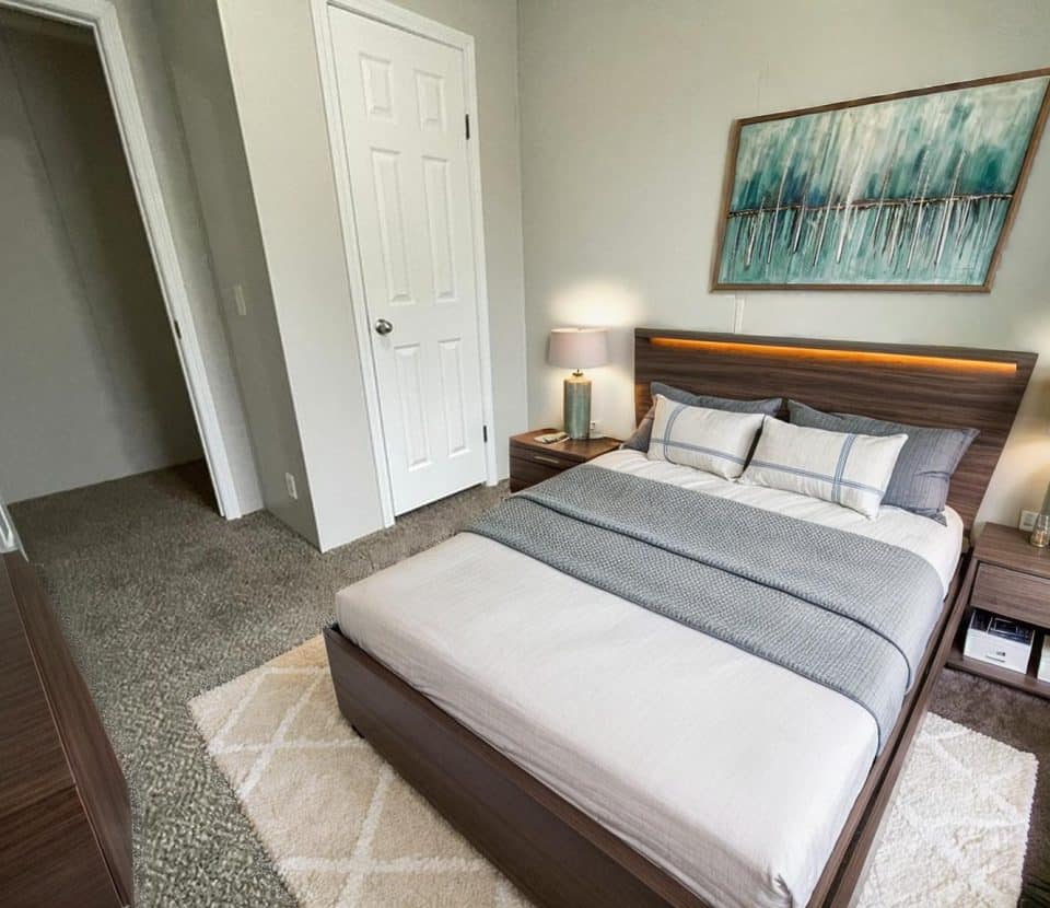A modern bedroom with a neatly made bed, two bedside tables with lamps, a large abstract painting above the headboard, and gray carpeted flooring. A white door is open, leading to a hallway.