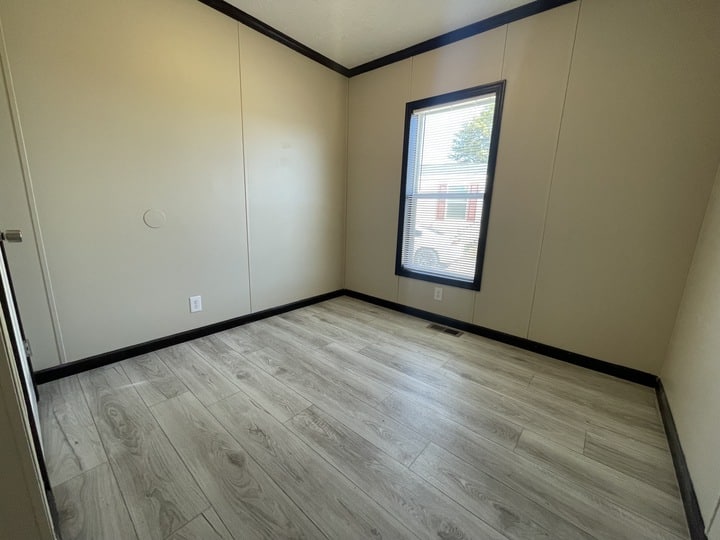 A small, empty room with light wood flooring, beige walls, a single window with blinds, and black trim along the ceiling and floor. Sunlight comes through the window, casting a shadow on the floor.
