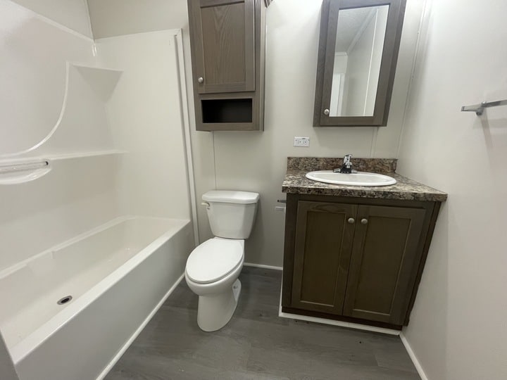 A small bathroom with a white bathtub, a toilet, a brown wood vanity with a sink, a matching wall cabinet, and light gray flooring. The walls are white and the countertop has a marble pattern.