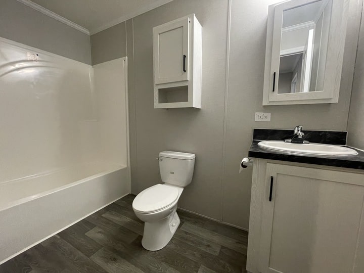 A modern bathroom with gray wood flooring, a white toilet, a white bathtub with a shower, a white vanity with a sink, and two white wall cabinets. The walls are light gray.