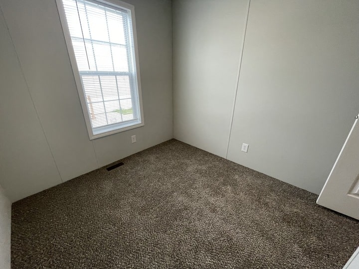 Small empty room with beige carpet, off-white walls, a closed white door, and a window letting in natural light. An electrical outlet and a floor vent are visible on the walls and floor.