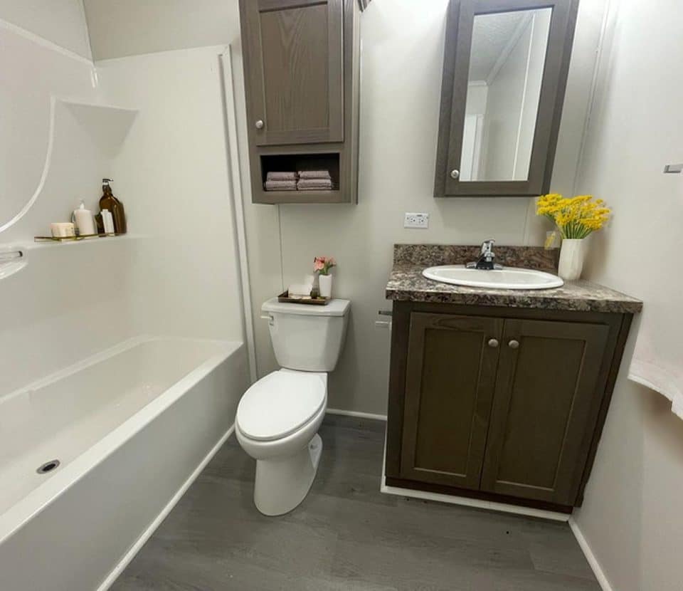 A modern bathroom with a bathtub, toilet, brown wood cabinets, a sink with a marble countertop, a mirror, white walls, gray flooring, and yellow flowers in a vase on the counter.