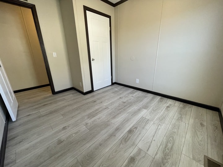 Empty room with light wood flooring, beige walls, white trim, and two white doors—one open and one closed. Minimalist and well-lit, with no furniture or decorations.
