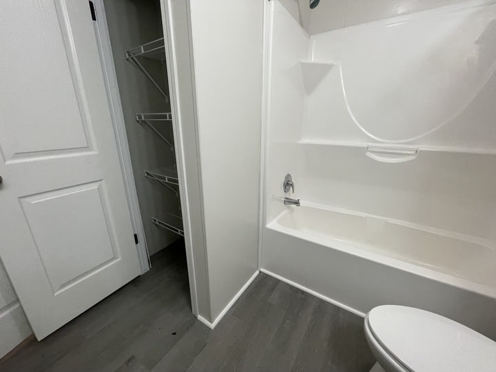 A bathroom with a white bathtub and shower, gray flooring, a toilet in the foreground, and an open closet with wire shelves next to a white door.