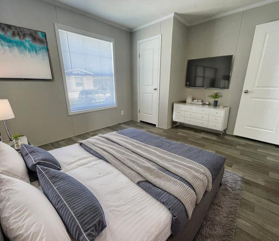 Modern bedroom with a neatly made bed in blue and white bedding, a nightstand with a lamp, a dresser with a TV, a potted plant, a large window with blinds, wood flooring, and minimal decor.