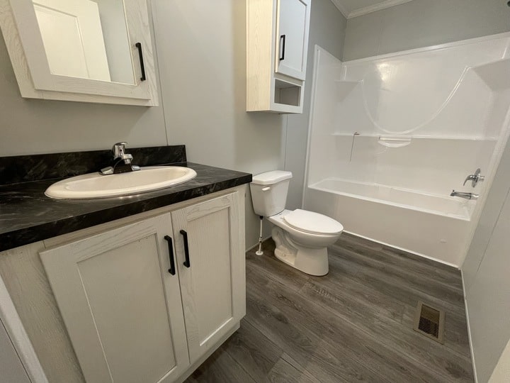 A modern bathroom with a white sink and vanity, toilet, bathtub-shower combo, white cabinets, and gray wood-look flooring. The walls are light-colored, and the room appears clean and well-lit.