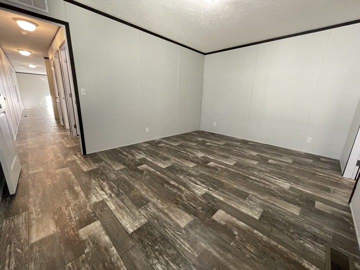 A spacious, empty room with light gray walls, dark trim, and wood-patterned vinyl flooring. A hallway with several doors and ceiling lights is visible to the left. The room is well-lit with natural and ceiling light.