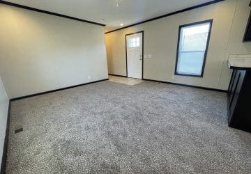 A spacious, empty living room with light-colored carpet, cream walls, black trim, two windows with blinds, and a white front door with a glass panel. The room is well-lit by natural light.