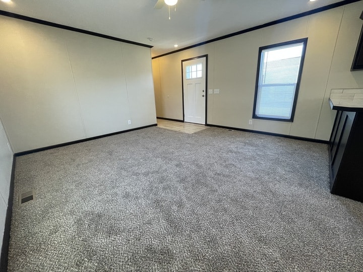 A spacious, empty living room with light-colored carpet, cream walls, black trim, two windows with blinds, and a white front door with a glass panel. The room is well-lit by natural light.