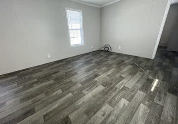Empty room with light gray walls, a single window with blinds, dark wood-patterned vinyl flooring, electrical outlets, and a coiled cable on the floor near the corner.