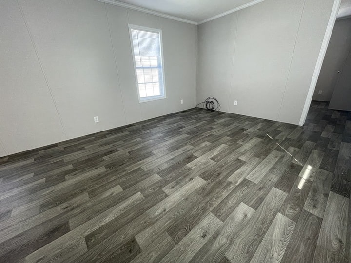 Empty room with light gray walls, a single window with blinds, dark wood-patterned vinyl flooring, electrical outlets, and a coiled cable on the floor near the corner.