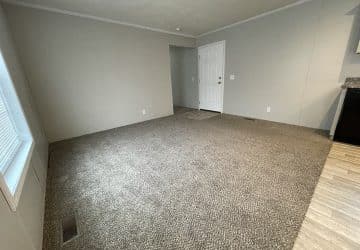 A spacious, empty room with light gray carpet, beige walls, a white door, and a section of light wood-look flooring near the door. A large window allows natural light into the room.