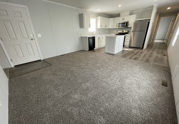 Open-concept living area with gray carpet, a modern kitchen with white cabinets, stainless steel appliances, an island, and light gray walls. A hallway leads to other rooms.