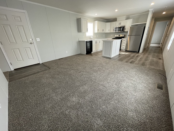 Open-concept living area with gray carpet, a modern kitchen with white cabinets, stainless steel appliances, an island, and light gray walls. A hallway leads to other rooms.