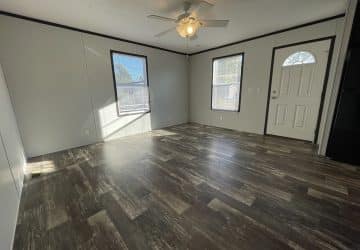 Bright, empty room with large windows, a ceiling fan, light gray walls, and dark wood-style flooring. A white door with a window is on the right side near a black kitchen appliance. Sunlight creates patterns on the floor.