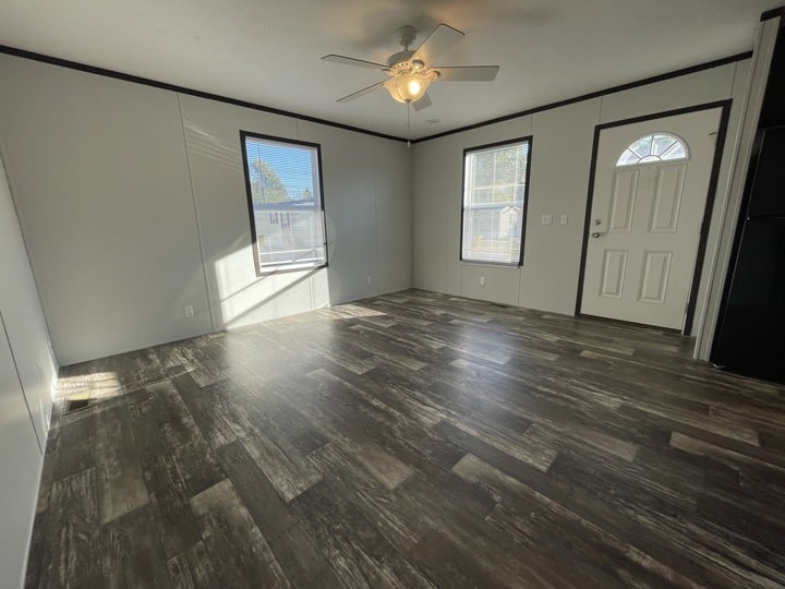 Bright, empty room with large windows, a ceiling fan, light gray walls, and dark wood-style flooring. A white door with a window is on the right side near a black kitchen appliance. Sunlight creates patterns on the floor.