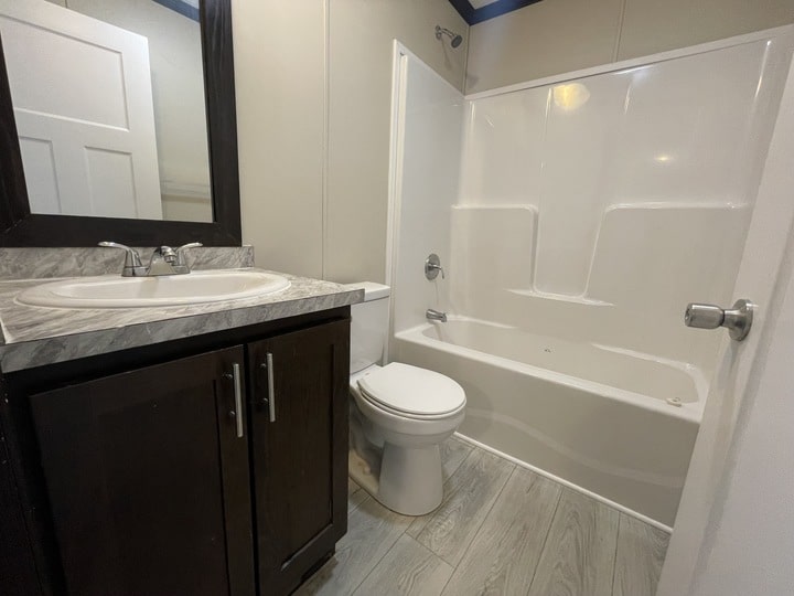 A small bathroom with a white bathtub and shower, a toilet, and a vanity with a marble countertop, sink, and dark wood cabinet. The floor has light wood-style tiles.