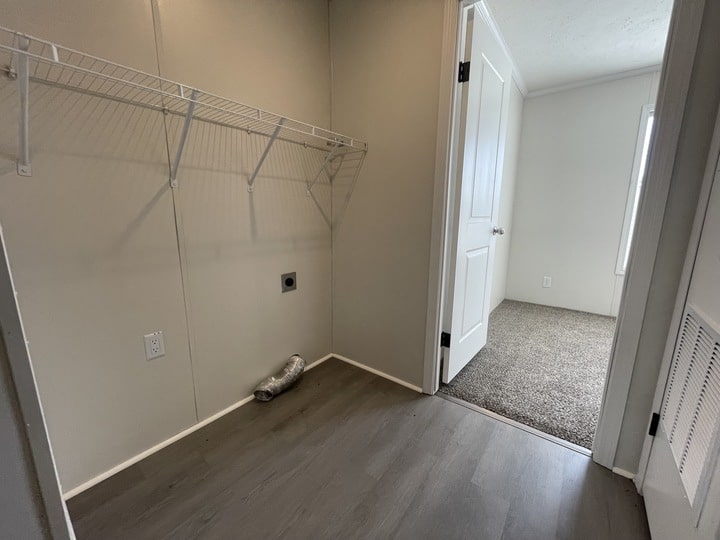Empty laundry room with gray flooring, a wire shelf on the left wall, dryer vent hose on the floor, and an open door leading to a carpeted bedroom. Walls are light-colored and the space appears clean and bright.