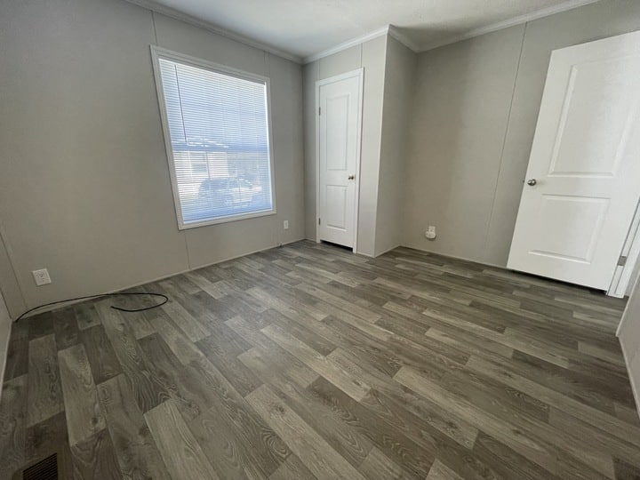 Empty room with gray wood-look flooring, light gray walls, a large window with blinds, and two white doors, one closed and one open. There is a black cable on the floor near the window.