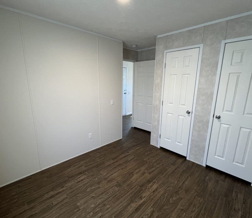 Small empty room with light-colored walls, two white closet doors, a white door leading to a hallway, and dark wood laminate flooring. The room is well-lit by natural light.