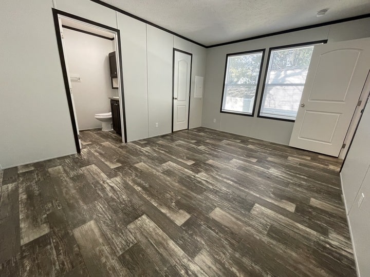 Empty room with wood-style flooring, white walls, large windows letting in natural light, an open door, and a view into a small bathroom with a toilet and dark vanity.