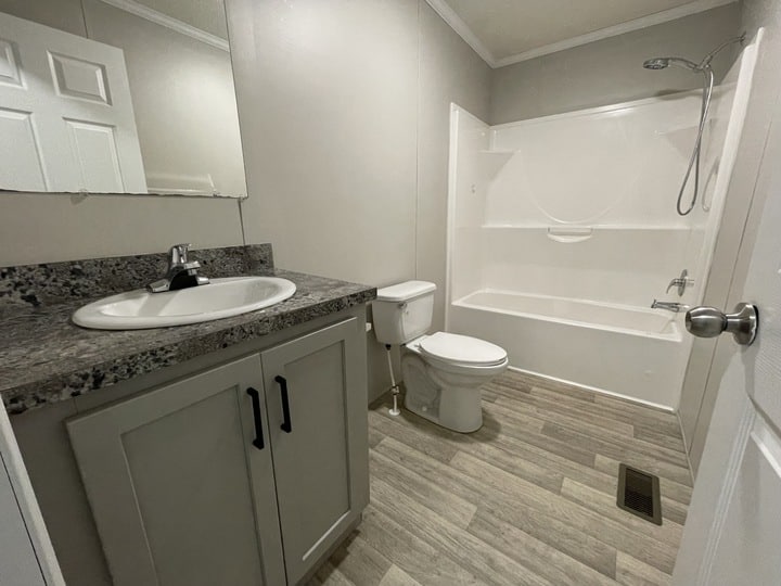 Modern bathroom with a granite countertop sink, white toilet, bathtub with shower, light wood-look flooring, and light gray walls. A mirror is above the sink, and the room has minimal decor.