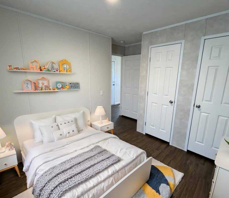 A bright and tidy bedroom with a white bed, bedside tables with lamps, two closed closets, wall shelves displaying decorative items, and a geometric rug on a dark wood floor.