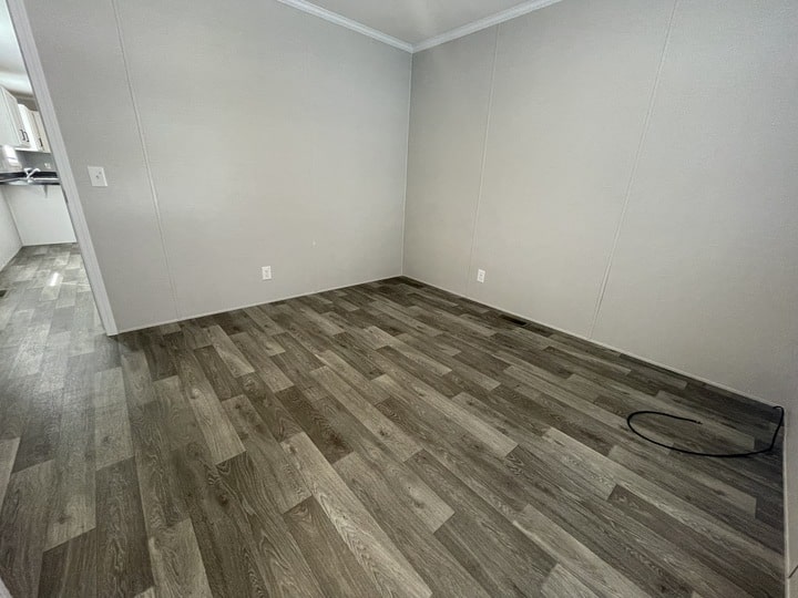 Empty room with light gray walls and wood-look vinyl flooring; a black cable lies on the floor near one corner. An open doorway leads to a kitchen area in the background.