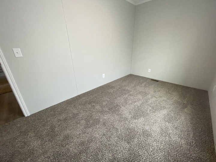An empty room with light gray carpet, light gray walls, white electrical outlets, and a white light switch. There is a vent on the floor and no furniture or decorations.