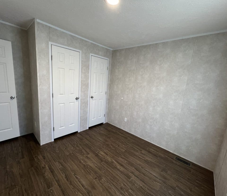 Empty room with light beige walls, dark wood flooring, a ceiling light, two closed white closet doors, one regular door, and a window letting in natural light on the right.