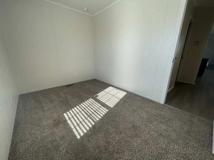 A small, empty room with light gray carpet, white walls, and sunlight streaming through window blinds, casting a shadow on the floor. An open doorway leads to a hallway with wood flooring.
