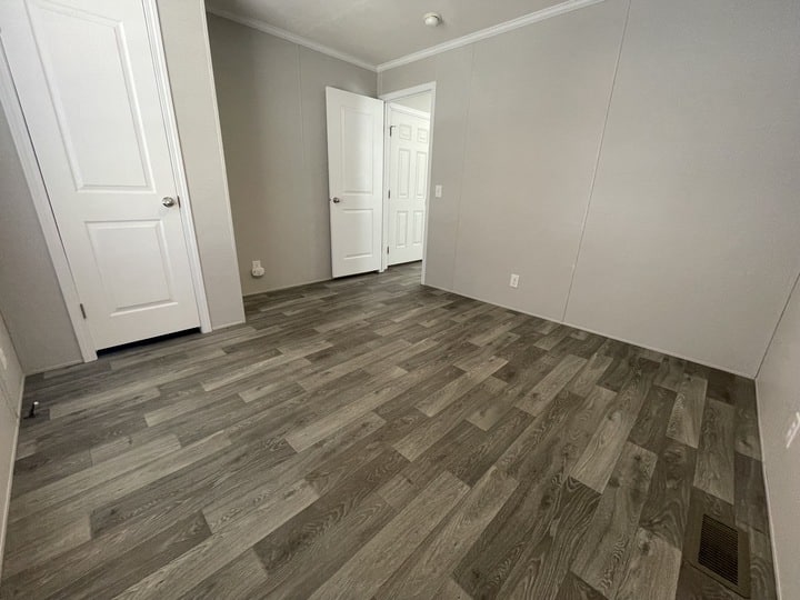 Empty room with gray wood-look flooring, light gray walls, and white trim. Two white doors are visible, one open and one closed, with a vent on the floor and an electrical outlet on the wall.