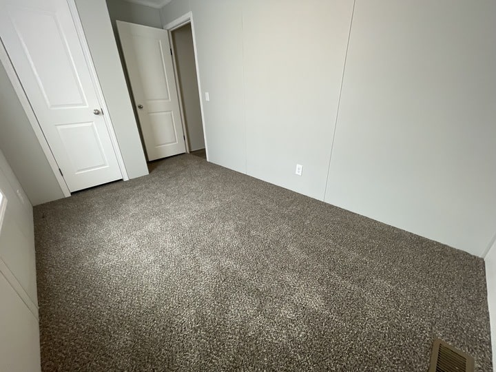 A small, empty room with light gray walls, two closed white doors, and brown carpeted flooring. There is a floor vent near the bottom right corner and natural light coming from the left.