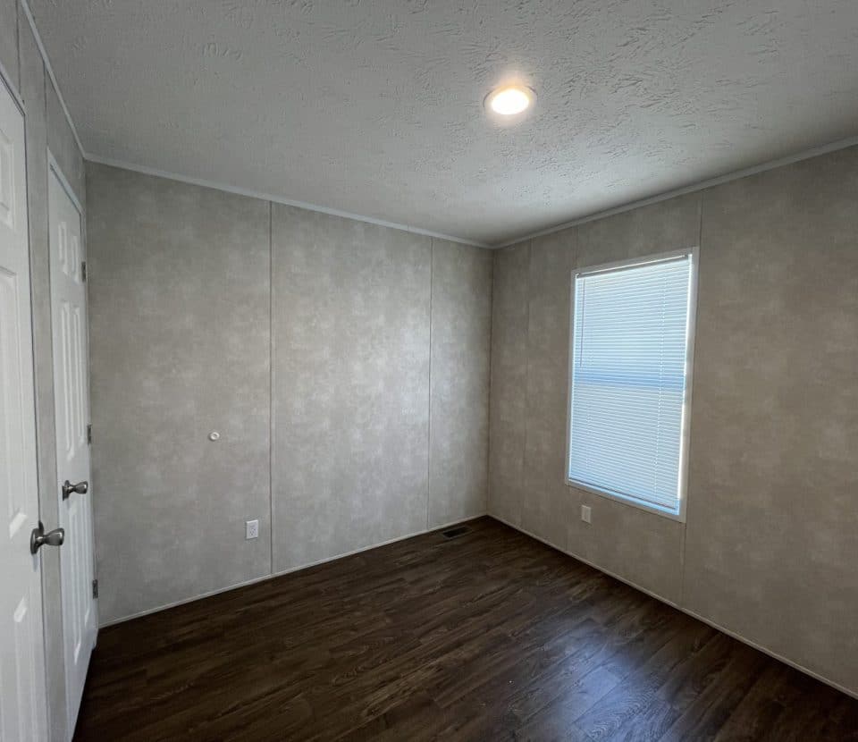 An empty room with light beige textured walls, dark wood flooring, a window with closed blinds, a ceiling light, and double white closet doors.