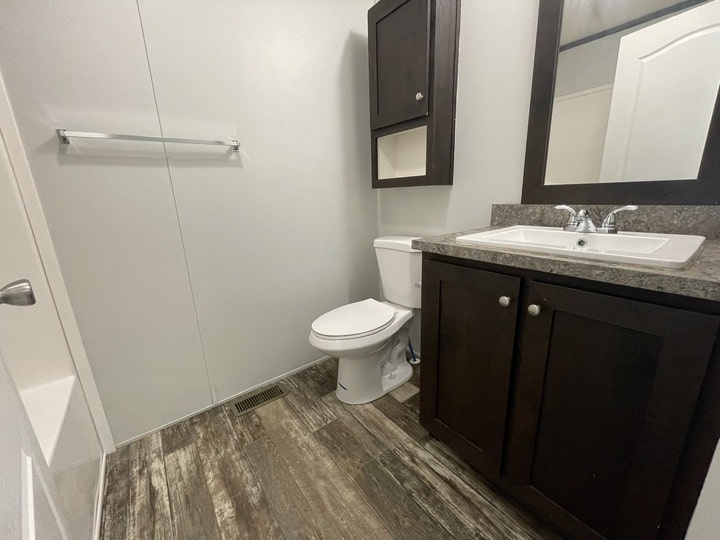 A small bathroom with wood-look flooring, a white toilet, a dark wood vanity and cabinet, a sink with a mirror above, and a towel bar on a plain wall next to a bathtub.