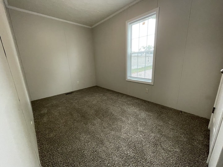 An empty room with beige walls, a carpeted floor, a single window letting in daylight, and a white door slightly open. The room has no furniture or decorations.
