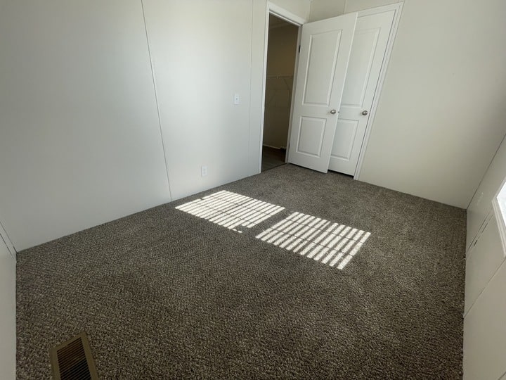 A small, empty room with light-colored walls, gray carpet, an open white door, and sunlight streaming through blinds, casting shadows on the floor.
