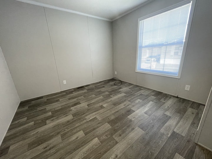 Empty room with grayish wood-look flooring, light gray walls, white trim, and a large window with blinds letting in natural light. An electrical outlet and a cable are visible near the floor.