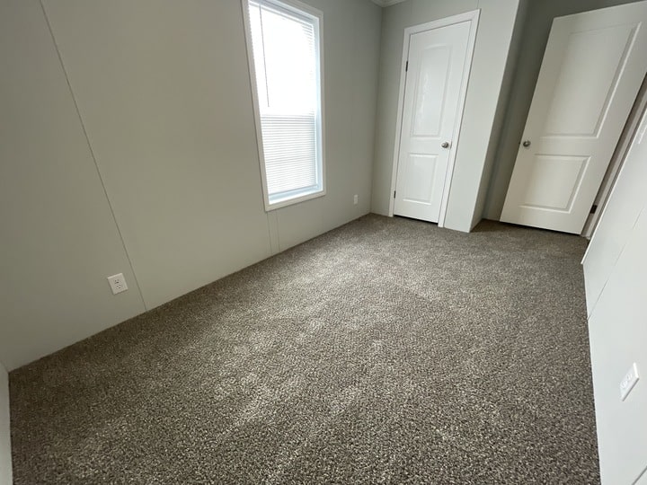 Empty room with light gray walls, beige carpet, a window with blinds on the left wall, and two white doors on the right wall, one closed and one partially open.