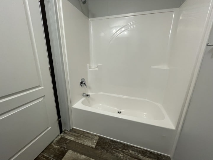 A clean, white bathtub and shower combination with a chrome faucet, surrounded by smooth white walls. The bathroom has light-colored wood-style flooring and a white door nearby.