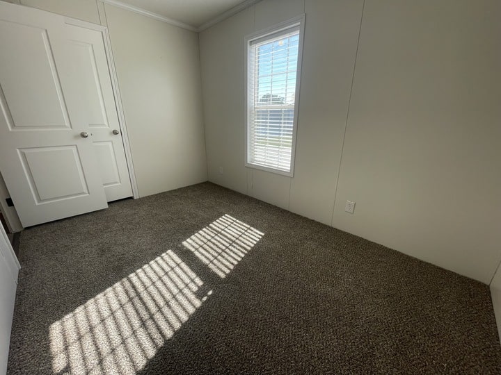 A small, empty room with beige walls, a closed white door, a window with blinds letting in sunlight, and brown carpeted floor. Shadows from the window are cast on the carpet.
