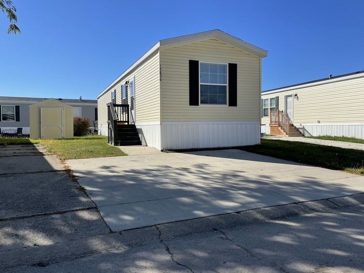 A beige mobile home with black shutters sits on a concrete driveway with a small grassy yard, next to similar homes, under a clear blue sky.