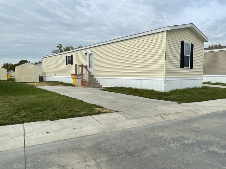 A beige manufactured home with white trim and black shutters sits on a grassy lot. A small set of stairs leads to the front door, and a concrete driveway is beside the home. A yellow shed is in the background.